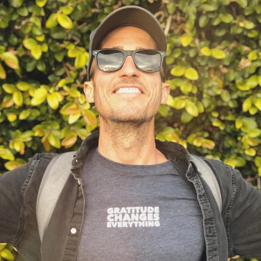 Aaron Lahman outdoors in a cap and sunglasses, smiling in front of a sunlit green hedge.