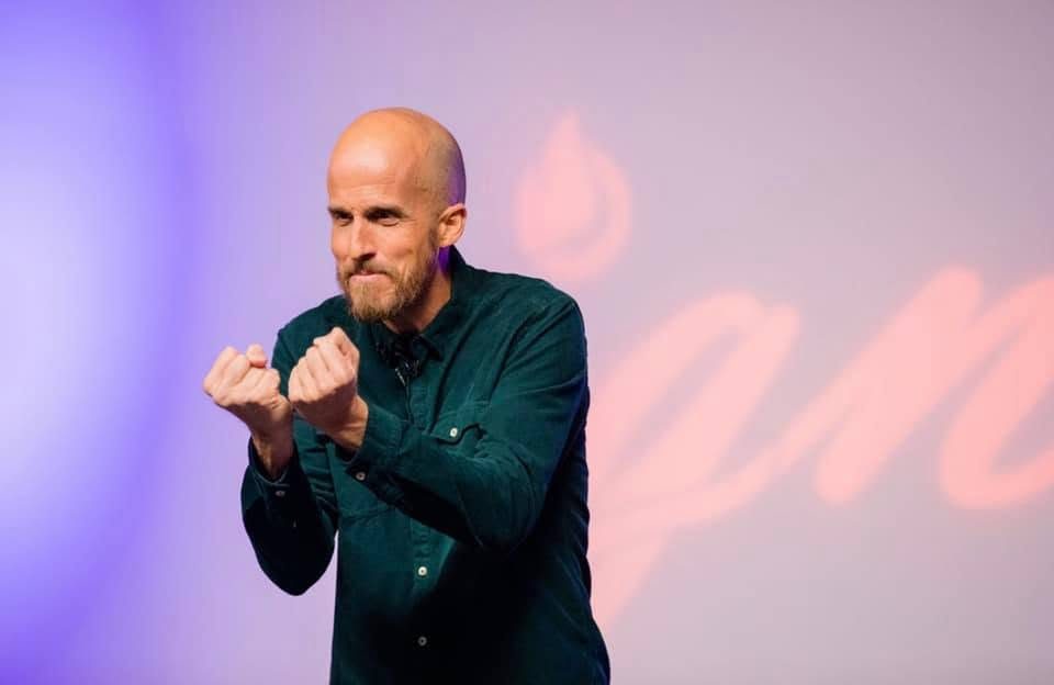 Aaron Lahman on stage in a dark green shirt, hands raised mid-gesture as he tells a story.
